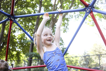 Glückliches süßes Mädchen klettert Klettergerüst gegen Bäume auf dem Spielplatz - CAVF54702