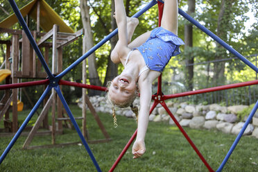 Porträt eines niedlichen glücklichen Mädchens, das kopfüber an einem Klettergerüst gegen Bäume auf einem Spielplatz hängt - CAVF54704