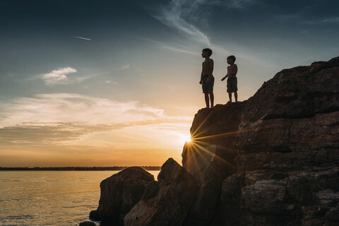 Low angle view of shirtless brothers standing on rocks by river against sky during sunset stock photo