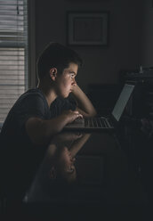 Side view of serious boy using laptop computer in darkroom - CAVF54749