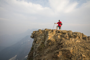 Wanderer in voller Länge mit Wanderstöcken auf einer Klippe stehend vor Bergen und bewölktem Himmel bei nebligem Wetter - CAVF54772