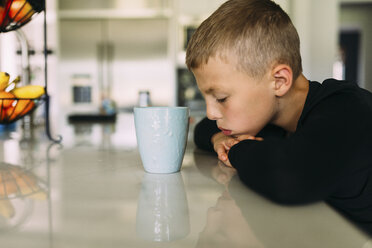 Seitenansicht eines müden Jungen mit einer Teetasse, der seinen Kopf auf den Küchentisch legt - CAVF54779