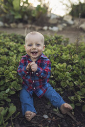 Porträt eines fröhlichen kleinen Jungen, der inmitten von Pflanzen im Park sitzt - CAVF54785