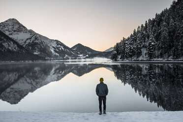 Rear view of man standing at lakeshore against sky during winter - CAVF54843