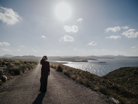 Frau geht auf der Straße am Meer gegen den Himmel an einem sonnigen Tag, lizenzfreies Stockfoto