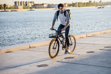 Young man with backpack riding bike on waterfront promenade at the riverside - VPIF01055