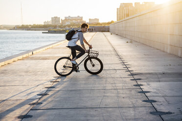 Young man with backpack riding bike on waterfront promenade at the riverside at sunset - VPIF01057