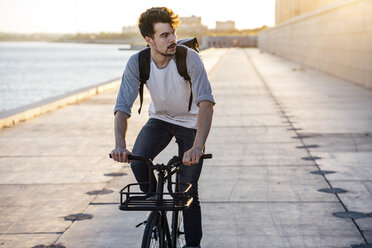 Young man with backpack riding bike on waterfront promenade at the riverside - VPIF01058