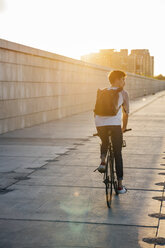 Young man with backpack riding bike on promenade at sunset - VPIF01059