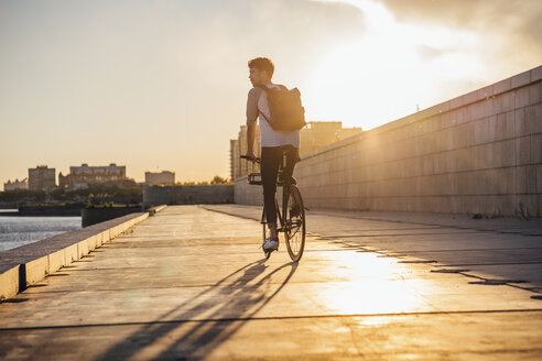 Junger Mann mit Rucksack fährt Fahrrad auf der Uferpromenade am Flussufer bei Sonnenuntergang - VPIF01060