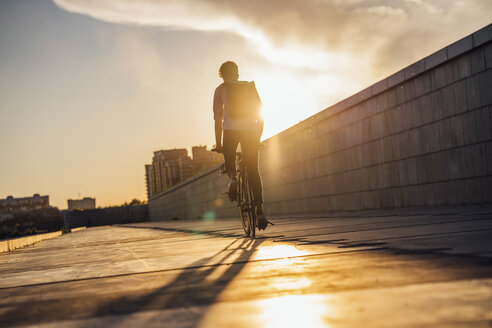 Junger Mann mit Rucksack fährt Fahrrad auf der Promenade bei Sonnenuntergang - VPIF01063