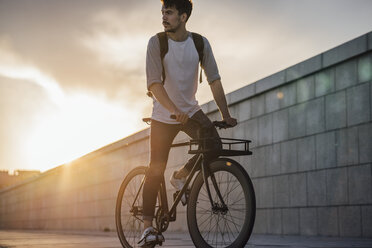 Young man with backpack riding bike on promenade at sunset - VPIF01064