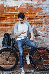 Young man with commuter fixie bike standing at brick wall with cell phone and earphones - VPIF01087