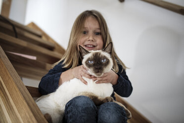 Little girl sitting on stairs with her Siamese cat at home - KMKF00639