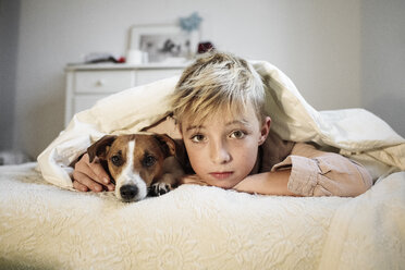 Porträt eines blonden Jungen und seines Jack Russel Terriers, die zusammen auf einem Bett liegen - KMKF00648