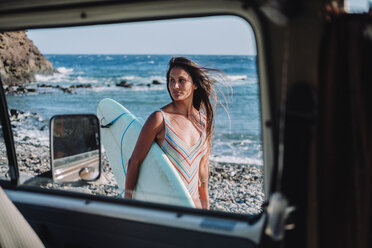 View from inside camper van of female surfer walking with surfboard on stony beach by sea, Tenerife, Canary Islands, Spain - AURF07787