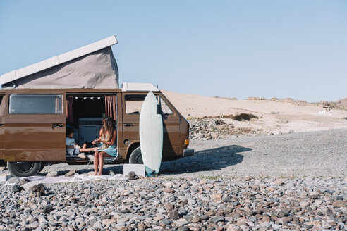 Pulled back view of young family sitting by vintage camper van on cobblestone beach, Tenerife, Canary Islands, Spain - AURF07790