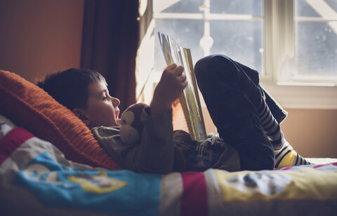 Junge mit Plüschtier, der zu Hause auf dem Bett liegt und ein Buch liest, lizenzfreies Stockfoto