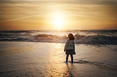 Rear view of girl standing on shore at beach against sky during sunset - CAVF55021