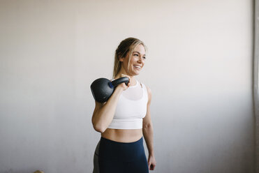 Lächelnde Athletin, die eine Kettlebell trägt, während sie an einer Wand im Fitnessstudio steht - CAVF55027