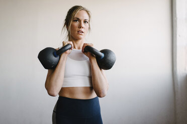 Porträt einer Athletin, die Kettlebells trägt, während sie an einer Wand im Fitnessstudio steht - CAVF55028