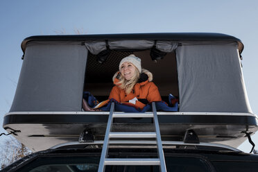 Niedriger Blickwinkel auf eine lächelnde Frau, die in einem Dachzelt auf einem Auto sitzt - CAVF55056