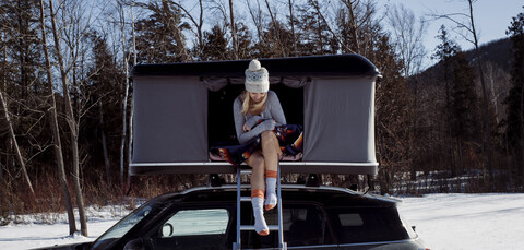 Frau in voller Länge im Dachzelt auf dem Auto sitzend im Winter, lizenzfreies Stockfoto