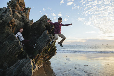 Playful boy jumping at beach while brother sitting on rocks - CAVF55082