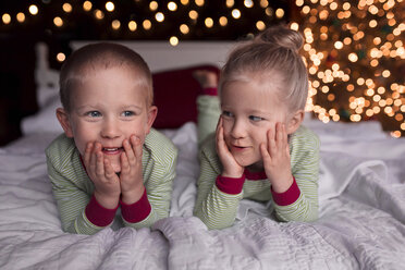 Geschwister, die sich auf dem Bett liegend vor dem beleuchteten Weihnachtsbaum zu Hause berühren - CAVF55174