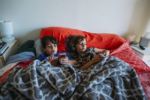 Hochformatige Ansicht von Geschwistern, die auf dem Bett liegend Smartphones benutzen, lizenzfreies Stockfoto