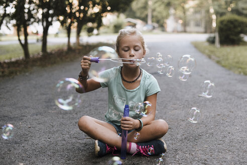 Girl with cross-legged blowing bubbles while sitting on road - CAVF55237