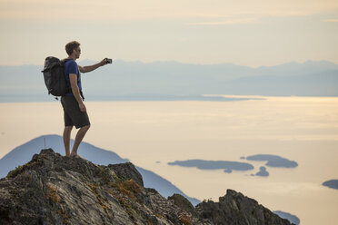 Seitenansicht eines Wanderers mit Rucksack, der auf einem Berg stehend gegen den Himmel fotografiert - CAVF55252