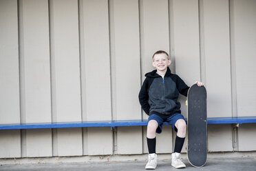 Porträt eines lächelnden Jungen, der ein Skateboard hält und auf einer Bank an der Wand sitzt - CAVF55352