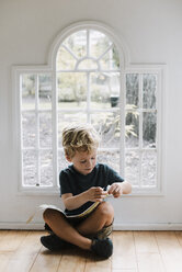 Boy with book sitting on floor against window at home - CAVF55355