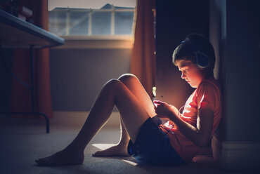 Side view of boy listening music while sitting on floor in darkroom at home - CAVF55357