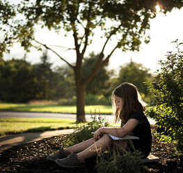 Side view of girl writing on book while sitting at park - CAVF55447