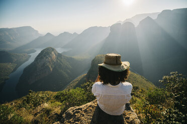 Rückansicht einer Frau mit Hut, die auf einem Berg sitzend die Aussicht gegen den Himmel an einem sonnigen Tag betrachtet - CAVF55467