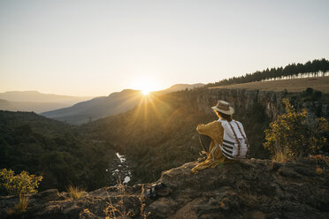 Side view of woman with backpack looking at view while sitting on mountain against sky during sunset - CAVF55468