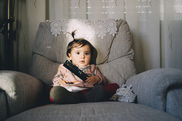 Niedliches Babymädchen mit Fernbedienung, das zu Hause auf einem Sessel sitzt - CAVF55519