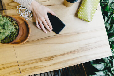 Abgeschnittene Hand einer jungen Frau, die ein Smartphone auf einem Holztisch in einem Café hält - CAVF55548