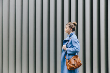 Seitenansicht einer jungen Frau, die einen Trenchcoat trägt und ihre Handtasche gegen einen Fensterladen in der Stadt hält - CAVF55551