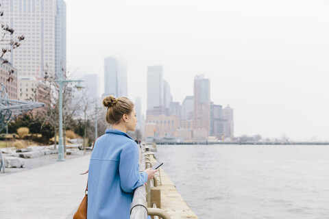 Junge Frau trägt Trenchcoat und schaut auf den Fluss an einem Geländer in der Stadt, lizenzfreies Stockfoto