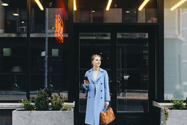 Junge Frau mit Smartphone und Handtasche, die einen Trenchcoat trägt, während sie vor dem Eingang eines Gebäudes in der Stadt steht - CAVF55566