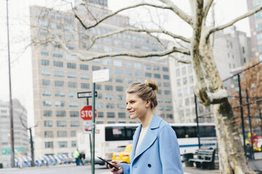 Seitenansicht einer lächelnden jungen Frau mit Smartphone, die auf der Straße vor einem Gebäude in der Stadt steht - CAVF55569