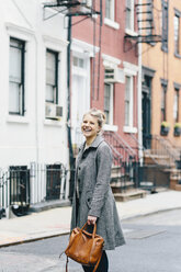Porträt einer fröhlichen jungen Frau, die einen Trenchcoat trägt und vor einem Wohnhaus in der Stadt steht - CAVF55575