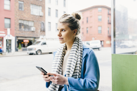 Junge Frau schaut weg, während sie ein Smartphone in der Stadt benutzt, lizenzfreies Stockfoto