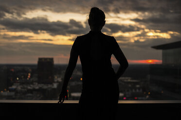 Rückansicht einer Frau, die auf einer Gebäudeterrasse steht, gegen die Stadt bei Sonnenuntergang - CAVF55594