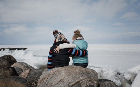 Rückansicht von Geschwistern, die auf einem Felsen am zugefrorenen See sitzen, gegen einen bewölkten Himmel, lizenzfreies Stockfoto