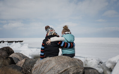 Rear view of siblings sitting on rock by frozen lake against cloudy sky - CAVF55601