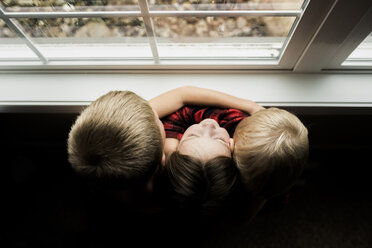 Hohe Winkelansicht von Geschwistern, die sich umarmen, während sie zu Hause am Fenster stehen - CAVF55620